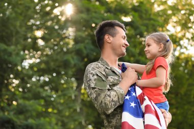 Amerikan bayrağı taşıyan askeri üniformalı bir baba kızını yeşil parkta tutuyor.