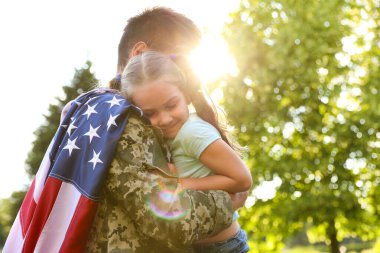 Amerikan bayrağı taşıyan askeri üniformalı bir baba kızını yeşil parkta tutuyor.