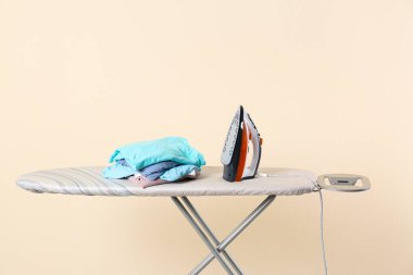 Board with modern iron and clean laundry on beige background