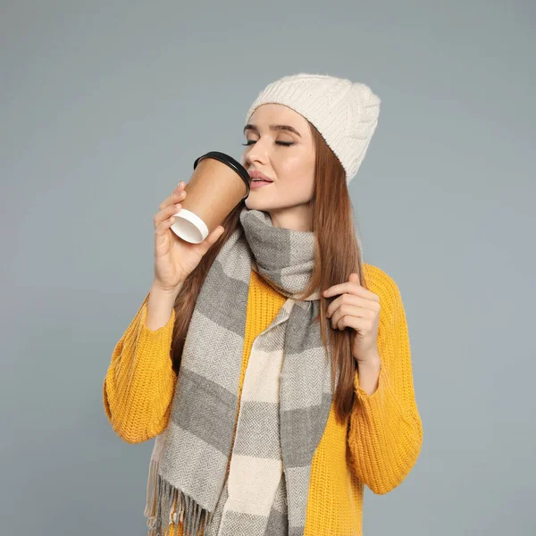 Šťastná mladá žena v teplém oblečení s pitím na šedém pozadí. Zimní sezóna — Stock fotografie