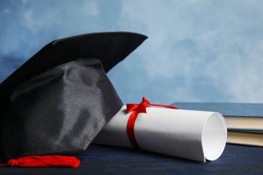 Mezuniyet şapkası, kitaplar ve öğrenci diploması açık mavi arka plana karşı koyu mavi ahşap masa