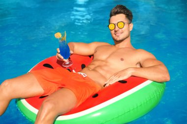 Güneşli bir günde yüzme havuzunda kokteyl ve şişme yüzük taşıyan genç bir adam. Yaz trendi olarak meyveli güneş gözlüğü