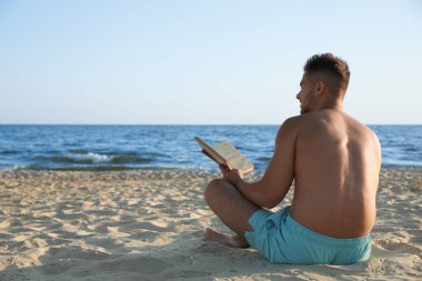 Genç adam deniz kenarındaki kumsalda kitap okuyor.