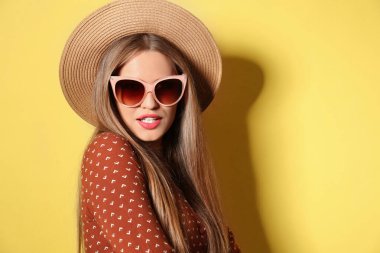 Sarı arka planda şık güneş gözlüğü ve şapka takan genç bir kadın.