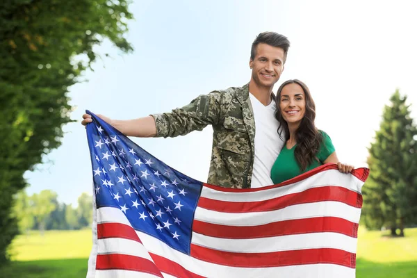 Homem de uniforme militar com bandeira americana e sua esposa no parque ensolarado — Fotografia de Stock