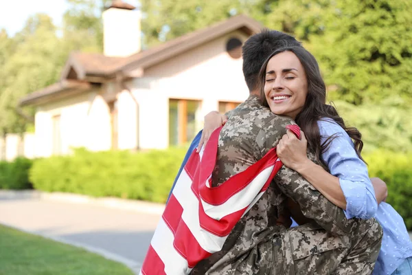 Hombre en uniforme militar con bandera americana abrazando a su esposa al aire libre — Foto de Stock