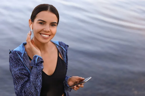 Joven deportista con auriculares inalámbricos y smartphone cerca del río — Foto de Stock