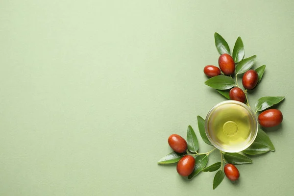 Glass bowl with jojoba oil and seeds on green background, flat lay. Space for text — Stock Photo, Image