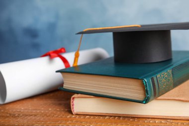 Mezuniyet şapkası, kitaplar ve öğrenci diploması açık mavi arka plana karşı ahşap masada, yakın plan.