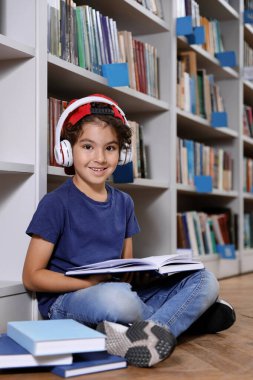 Kütüphanede yerde kitap okuyan kulaklıklı şirin bir çocuk.