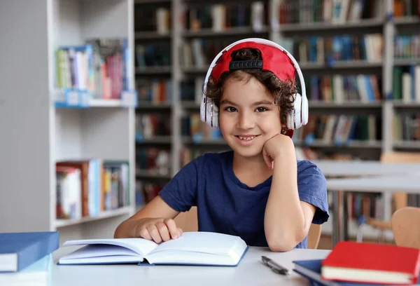 Lindo niño con auriculares leyendo libros en la mesa en la biblioteca — Foto de Stock