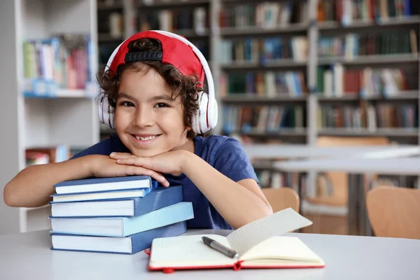 Lindo niño con auriculares y libros en la mesa en la sala de lectura de la biblioteca — Foto de Stock