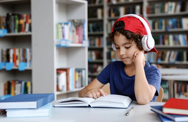 Симпатичный мальчик в наушниках, читающий книги за столом в библиотеке. — стоковое фото
