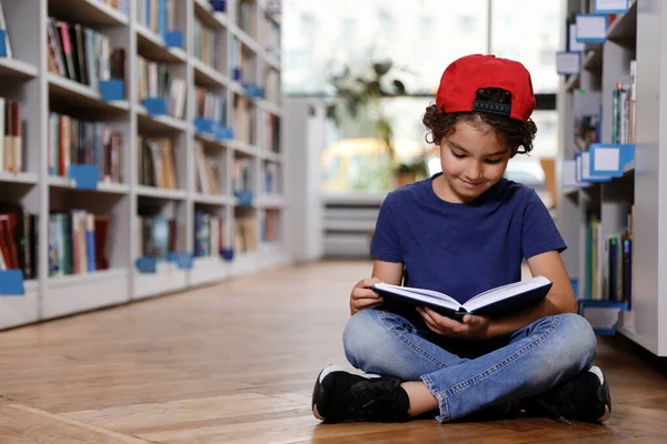 Lindo niño leyendo libro en el suelo en la biblioteca — Foto de Stock