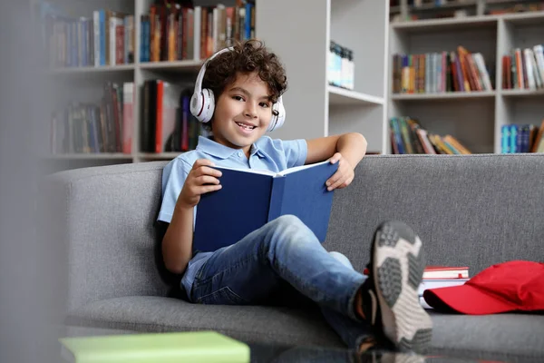 Lindo niño en auriculares libro de lectura en el sofá en la biblioteca — Foto de Stock