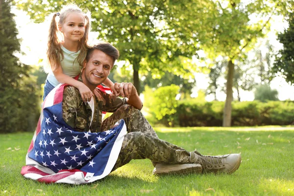 Padre en uniforme militar con bandera americana y su pequeña hija sentada en el césped en el parque — Foto de Stock