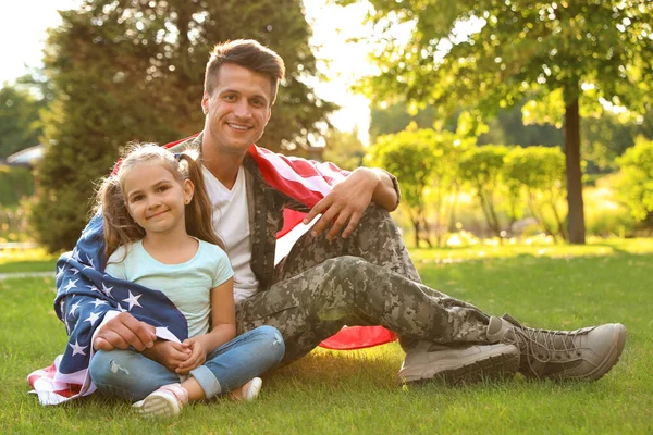 Pai de uniforme militar com bandeira americana e sua filhinha sentada na grama no parque — Fotografia de Stock