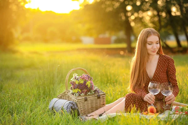 Gyönyörű fiatal nő borosüveggel és piknik kosárral ül a takarón a parkban — Stock Fotó