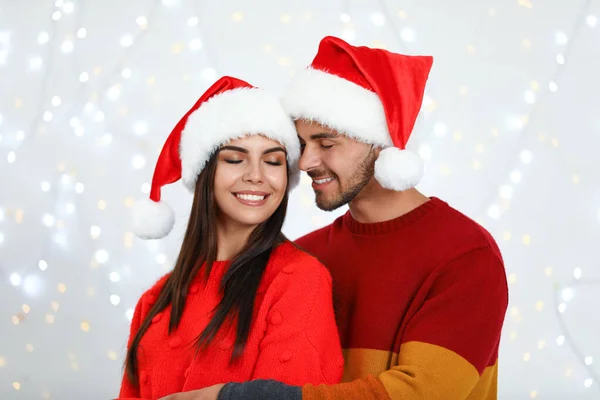 Preciosa pareja joven en los sombreros de Santa contra las luces borrosas festivas. Celebración de Navidad —  Fotos de Stock