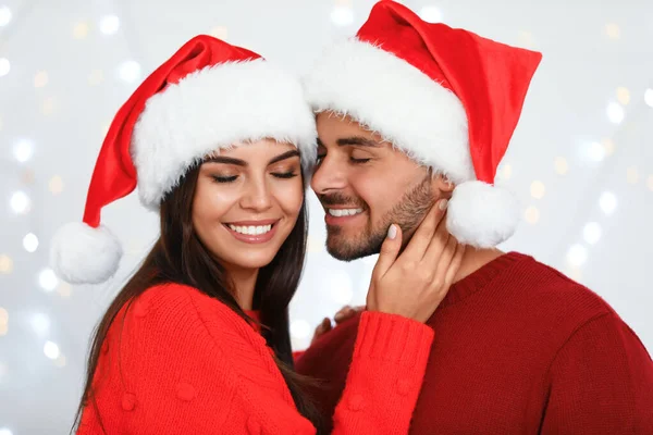 Lovely young couple in Santa hats against blurred festive lights. Christmas celebration — Stock Photo, Image