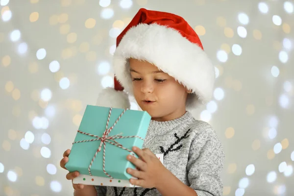 Удивленный маленький ребенок в шляпе Санты с подарочной коробкой против размытых праздничных огней. Праздник Рождества — стоковое фото