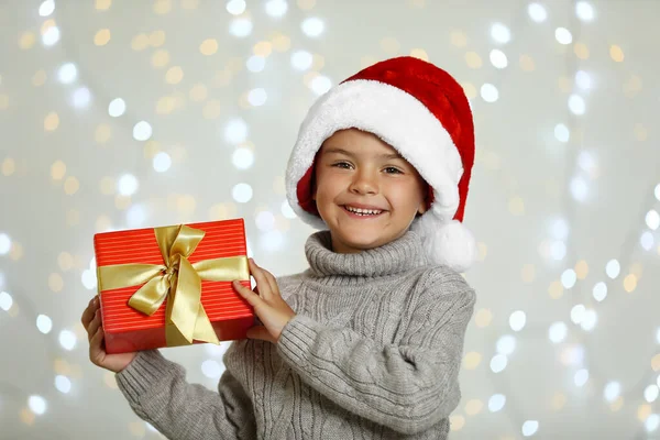 Criança feliz em chapéu de Papai Noel com caixa de presente contra luzes festivas borradas. Celebração de Natal — Fotografia de Stock