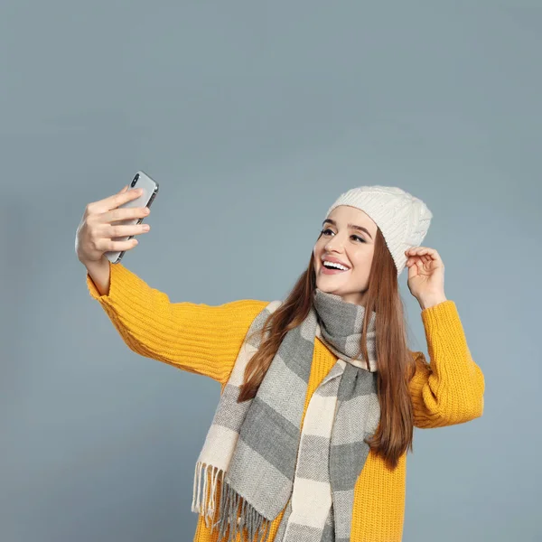 Όμορφη νεαρή γυναίκα με ζεστά ρούχα που βγάζει selfie σε γκρι φόντο. Χειμερινή περίοδος — Φωτογραφία Αρχείου
