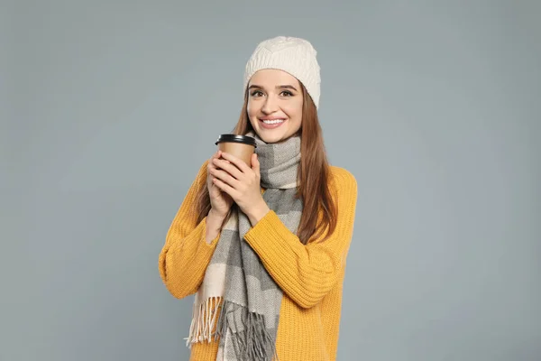 Счастливая молодая женщина в теплой одежде с напитком на сером фоне. Зимний сезон — стоковое фото