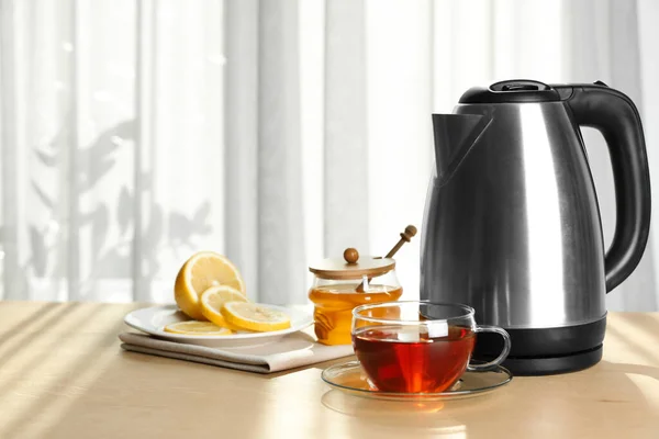 Современный электрический чайник и чашка чая на деревянном столе в помещении. Пространство для текста — стоковое фото