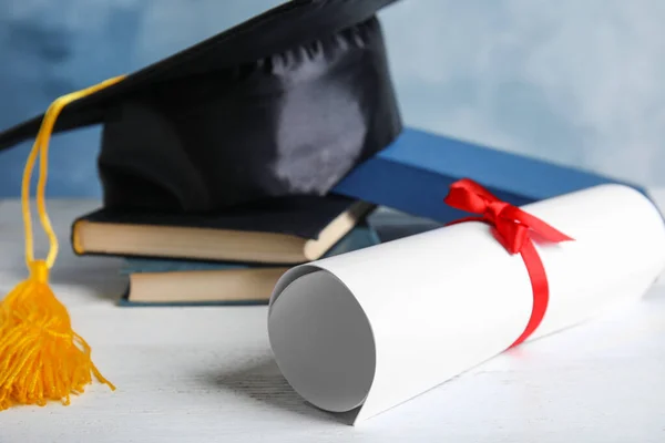 Sombrero de graduación, libros y diploma de estudiante en mesa de madera blanca sobre fondo azul claro, primer plano — Foto de Stock