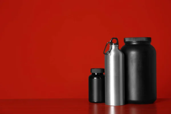 Frascos pretos com pó de proteína e garrafa na mesa contra fundo vermelho. Espaço para texto — Fotografia de Stock