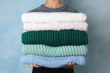 Woman holding stack of warm clothes on light blue background, closeup. Autumn season clipart