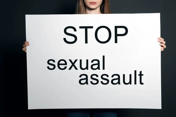 Young woman holding card with words STOP SEXUAL ASSAULT against dark background, closeup — Stock Photo, Image