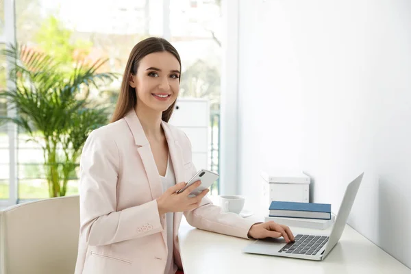 Młoda bizneswoman ze smartfonem za pomocą laptopa przy stole w biurze — Zdjęcie stockowe