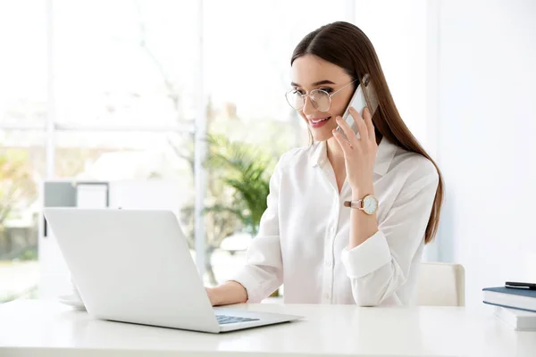 Jonge zakenvrouw praten over de telefoon tijdens het gebruik van laptop aan tafel in het kantoor — Stockfoto