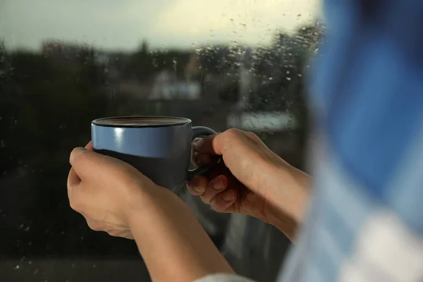 Jeune femme tenant une tasse de café à la fenêtre avec des gouttes de pluie, gros plan — Photo