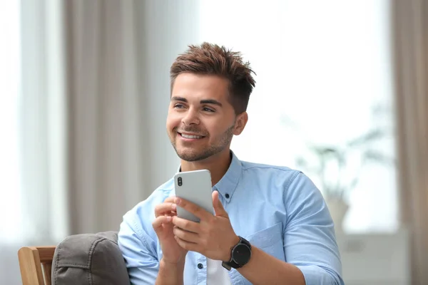 Retrato de joven guapo con teléfono inteligente sentado en la silla en la habitación — Foto de Stock