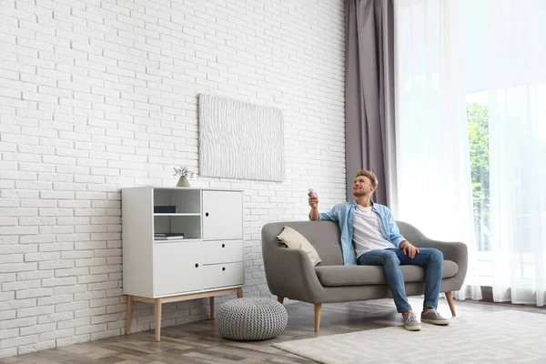 Happy young man switching on air conditioner on sofa at home
