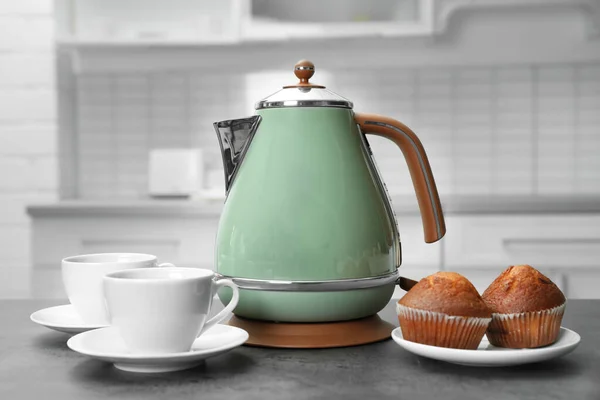 Modern electric kettle, cups and muffins on grey table in kitchen — Stock Photo, Image