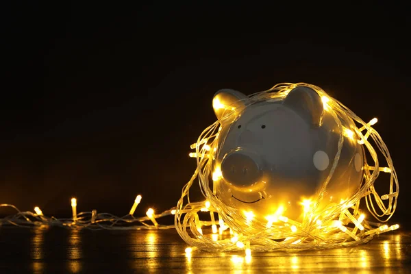 Banco porquinho com luzes de Natal na mesa de madeira contra fundo preto. Espaço para texto — Fotografia de Stock