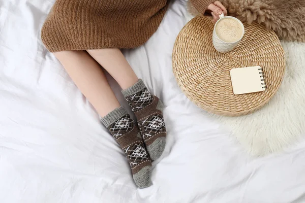 Woman with cup of hot drink wearing knitted socks on white fabric, top view. Warm clothes — Stockfoto