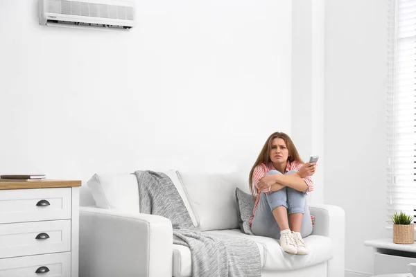 Young woman with air conditioner remote control freezing on sofa at home