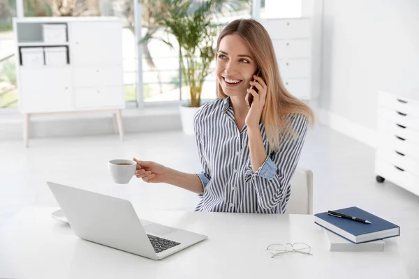 Joven mujer de negocios hablando por teléfono mientras usa el ordenador portátil en la mesa en la oficina — Foto de Stock