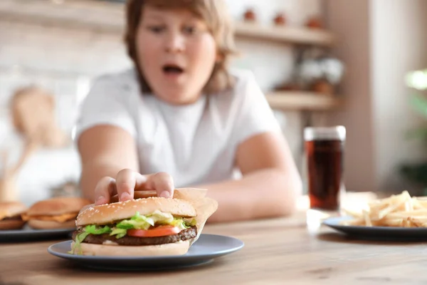 Emotionele overgewicht jongen reikt naar hamburger aan tafel in de keuken — Stockfoto
