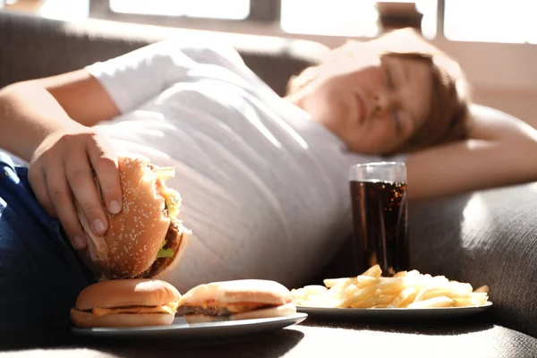 Ragazzo in sovrappeso dormire sul divano circondato da fast food — Foto Stock