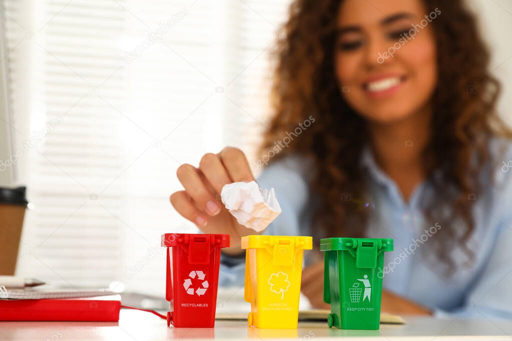 Young African-American woman throwing paper into mini recycling bin at table in office, focus on hand