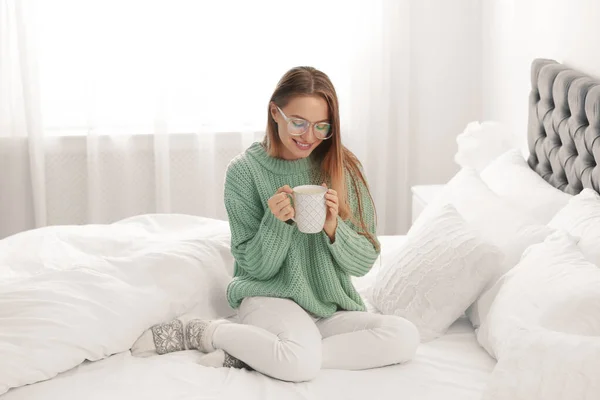 Mladá žena v teplém svetru s teplým nápojem na posteli doma — Stock fotografie