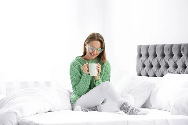 따뜻 한 스웨터를 입고 뜨거운 음료를 집 침대에 갖고 있는 젊은 여자 — 스톡 사진
