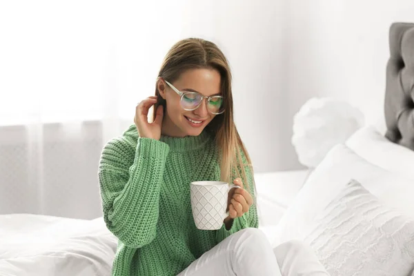 Młoda kobieta w ciepłym swetrze z gorącym napojem na łóżku w domu — Zdjęcie stockowe