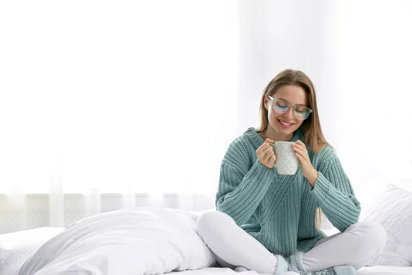 Giovane donna in maglione caldo con bevanda calda sul letto a casa. Spazio per testo — Foto Stock